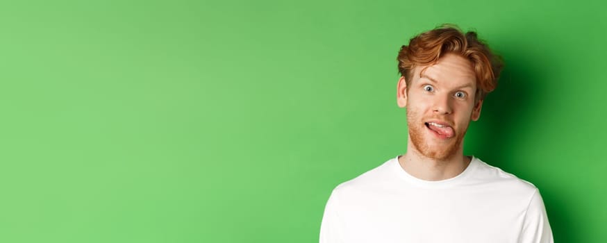 Emotions and fashion concept. Close up of funny redhead man showing silly faces, sticking tongue and staring at camera, standing over green background.