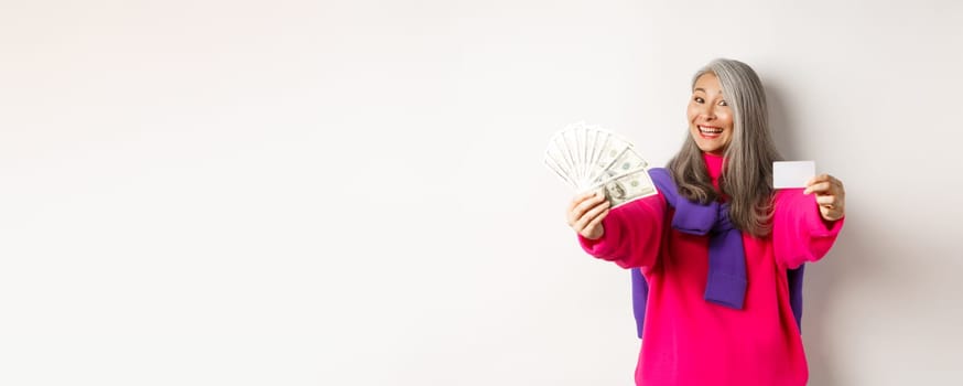 Shopping concept. Cheerful asian senior woman showing cash and plastic credit card, smiling at camera, standing over white background.