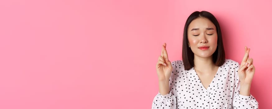 Beauty and lifestyle concept. Close-up of cute asian girl making a wish, close eyes and cross fingers hopeful, praying and standing over pink background.