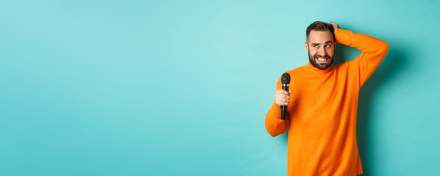 Shy and awkward adult man scratching head, holding microphone before singing, standing over blue background.