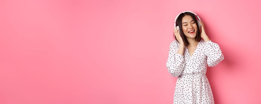 Romantic asian woman smiling happy, listening music in headphones and dancing, standing in trendy dress over pink background.