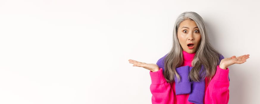 Close up of shocked asian mom staring at camera, spread hands sideways and gasping at camera confused, standing over white background.