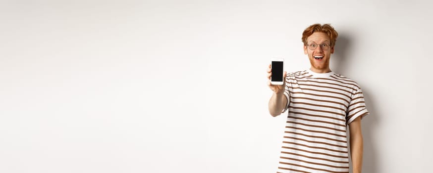 Technology and e-commerce concept. Happy young redhead man in glasses showing blank smartphone screen, looking at camera amazed, standing over white background.