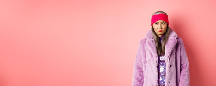 Sad and indecisive asian senior woman in fashionable faux fur coat looking left and sulking upset, standing against pink background.