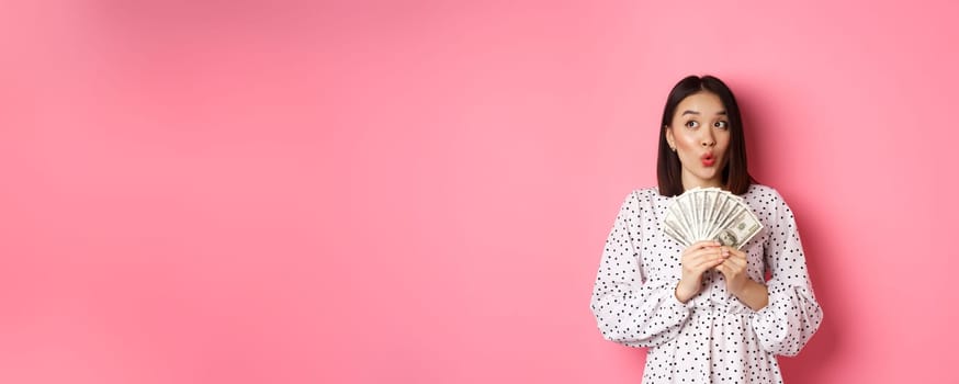 Shopping concept. Dreamy asian woman thinking, holding money dollars and looking aside thoughtful, standing over pink background.