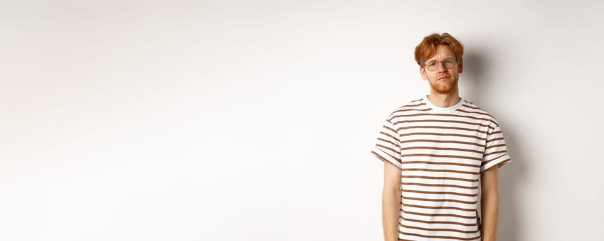 Reluctant and unamused redhead young man staring at camera, looking tired or displeased, standing over white background.