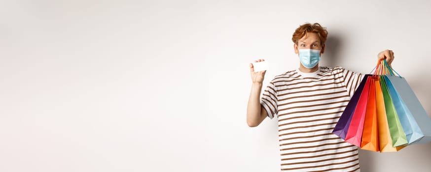 Social distancing, coronavirus concept. Young man with red hair, wearing face mask, showing shopping bags and plastic credit card, white background.