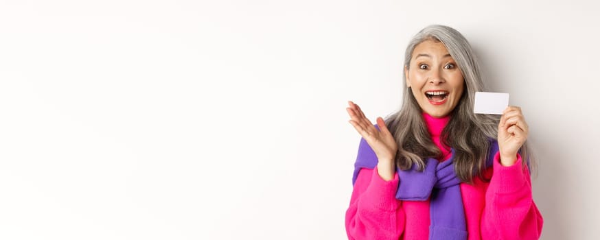 Shopping concept. Happy asian old aldy looking impressed and showing plastic credit card of her bank, standing over white background.