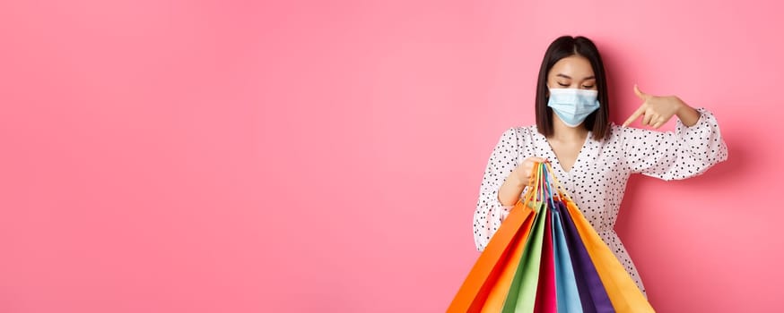 Covid-19, pandemic and lifestyle concept. Pretty korean woman in medical mask pointing at shopping bags, buying clothes, standing over pink background.