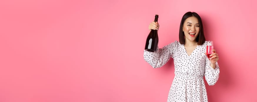 Happy asian woman celebrating, having fun and partying, pouring glass of champagne and laughing, standing over pink background.