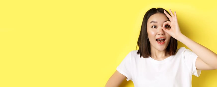Close up of cute asian girl feeling happy, showing OK sign on eye and smiling, standing over yellow background.