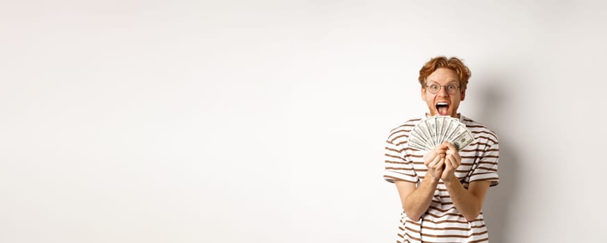 Lucky young man with red hair showing dollars, winning money and screaming of happiness, holding prize cash, standing over white background.