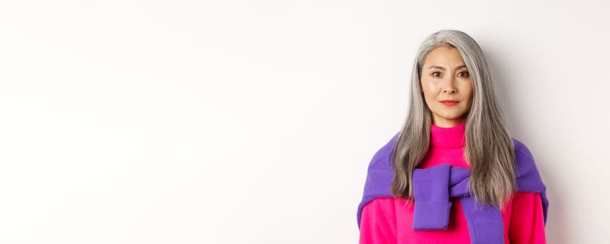 Close-up of stylish woman with grey hair and no wrinkles, smiling and looking at camera, standing in trendy outfit over white background.