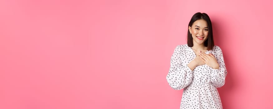 Thankful Korean girl in dress smiling, holding hands on heart and looking grateful at camera, touched with nice gesture, standing over pink background.