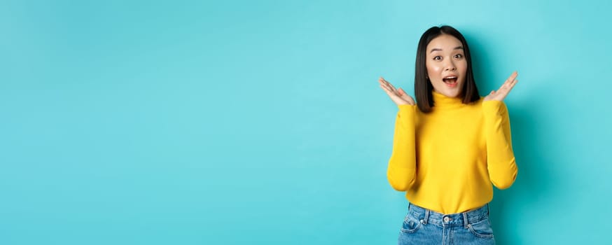 Beauty and fashion concept. Image of excited and surprised japanese girl saying wow with amazement, raising hands up near face, standing against blue background.