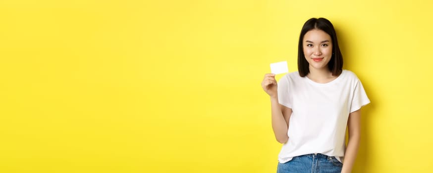 Young asian woman in casual white t-shirt showing plastic credit card and smiling at camera, yellow background.