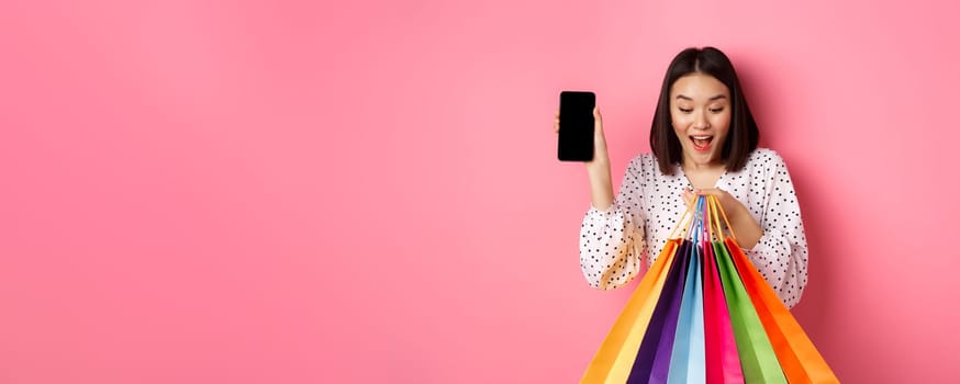 Attractive asian woman showing smartphone app and shopping bags, buying online via application, standing over pink background.