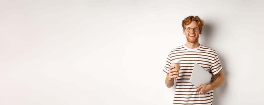 Handsome redhead male employee in glasses having break, drinking coffee and holding laptop, standing over white background.
