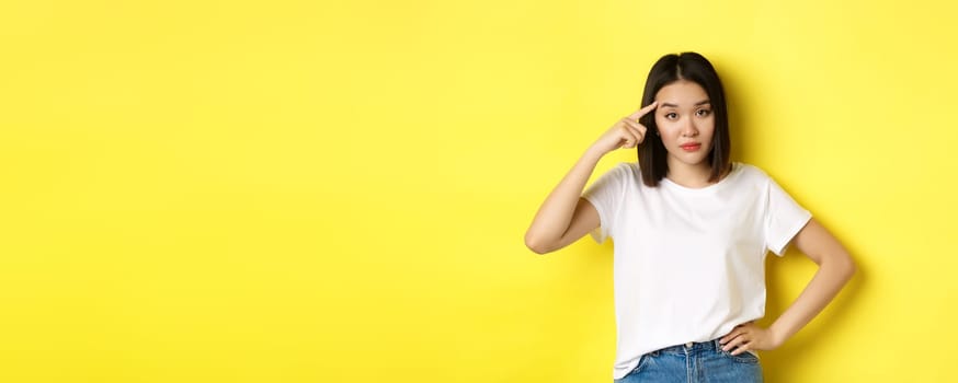 Annoyed young asian woman scolding someone stupid, pointing finger at head, asking are you crazy, standing over yellow background.