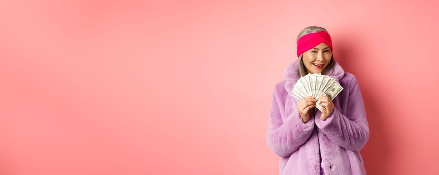 Shopping and fashion concept. Fashionable asian senior woman thinking about buying new clothes, showing money in dollars and smiling greedy, pink background.