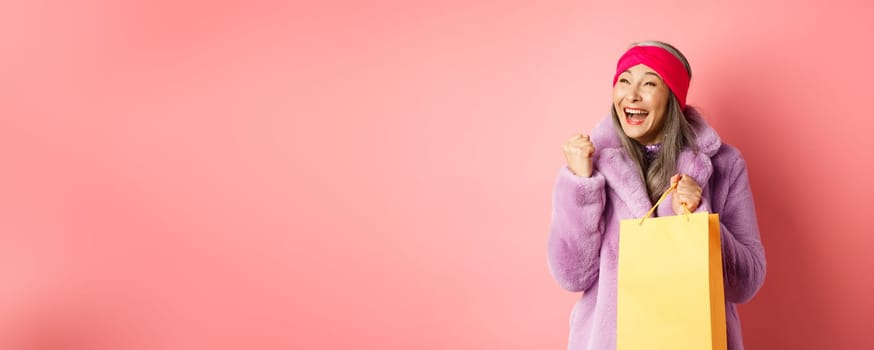 Shopping and fashion concept. Happy asian senior woman winning, holding paper bag and making fist pump gesture, saying yes with cheerful face, standing over pink background.