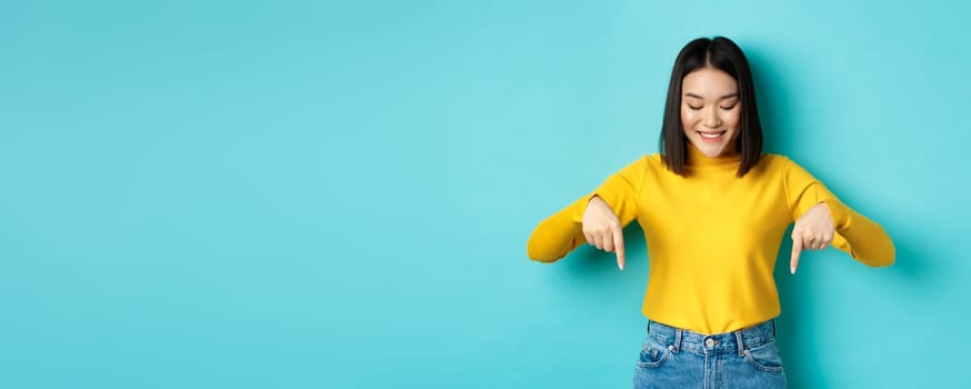 Shopping concept. Beautiful asian girl in yellow sweater smiling, pointing fingers down with satisfied face, showing cool advertisement, blue background.
