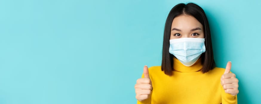 Covid-19, social distancing and pandemic concept. Close up of young asian woman in medical mask showing thumbs up, say yes, praise good offer, standing over blue background.