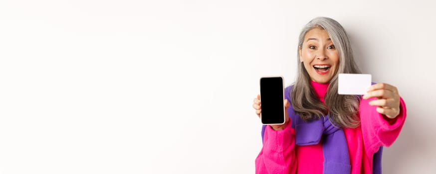 Online shopping. Close-up of stylish korean senior woman showing empty mobile screen and plastic credit card, smiling happy, standing over white background.