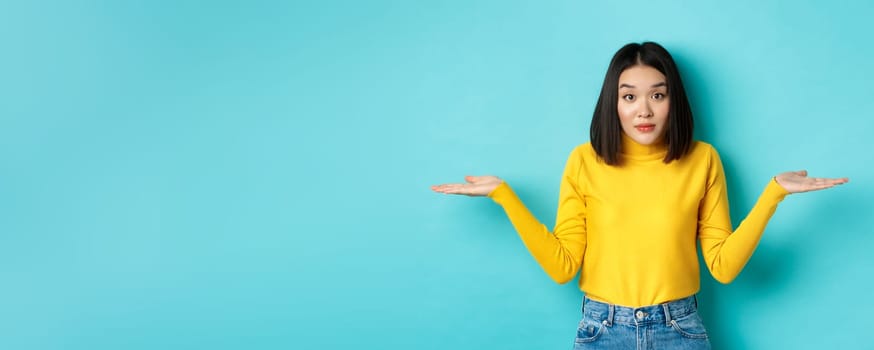 Image of indecisive asian woman shrugging shoulders, spread hands sideways and looking clueless at camera, standing indecisive against blue background.