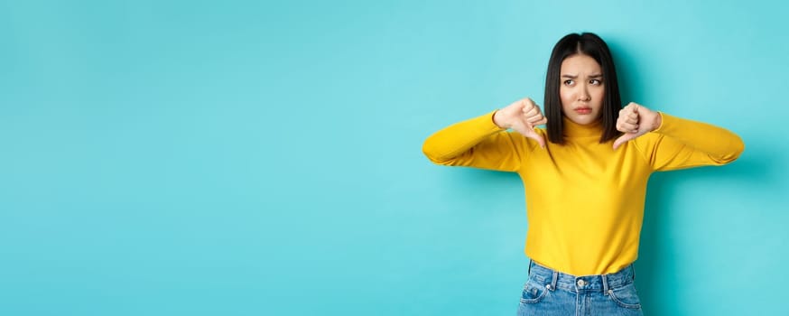 Disappointed asian woman frowning upset, showing thumbs down in dislike and disapproval, standing over blue background, looking left.