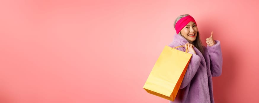 Stylish asian senior woman going shopping, carry paper bag on shoulder and showing thumb-up, recommending store discounts, pink background.