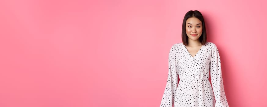 Beautiful asian lady smiling at camera, wearing cute romantic dress, standing over pink background. Copy space