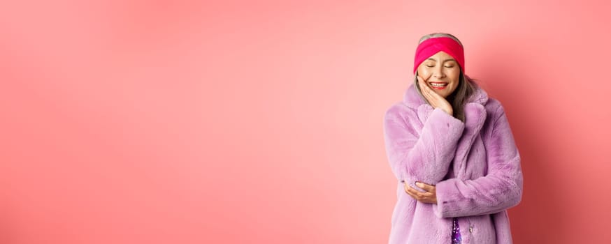 Fashion and shopping. Happy asian mature woman in purple faux fur coat smiling lovely and carefree, close eyes with happiness, standing against pink background.