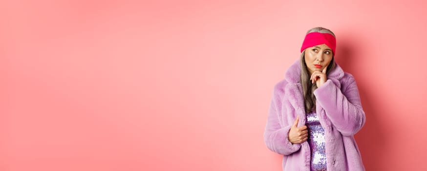 Fashionable asian senior woman looking doubtful, thinking and frowning indecisive, standing in purple faux fur coat and looking left, standing over pink background.