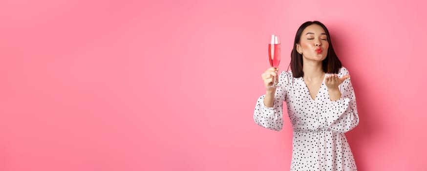 Romantic asian woman raising glass of champagne and sending air kiss at camera, celebrating and having fun, standing over pink background.