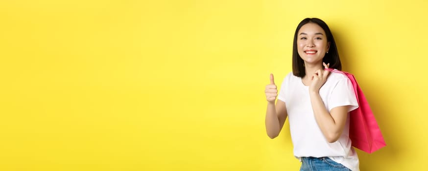 Cheerful asian female shopper looking amused, holding shopping bag and showing thumbs up, recommend store with discounts, standing over yellow background.