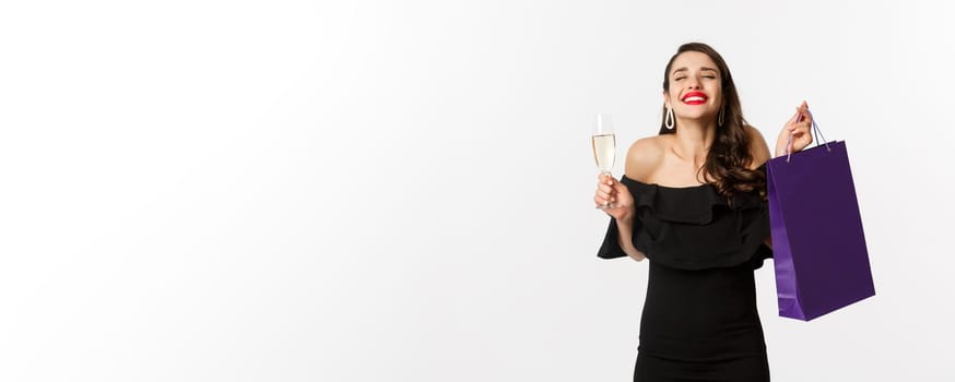Happy smiling woman celebrating, holding present in shopping bag and glass of champagne, standing in black dress over white background.