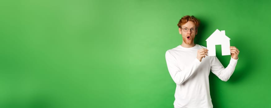 Real estate and buying property concept. Surprised young redhead man showing paper house model and looking amazed, standing over green background.