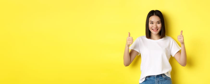 Pretty young asian woman in white t-shirt, showing thumbs up and smiling, praise good offer, recommend product, standing satisfied over yellow background.