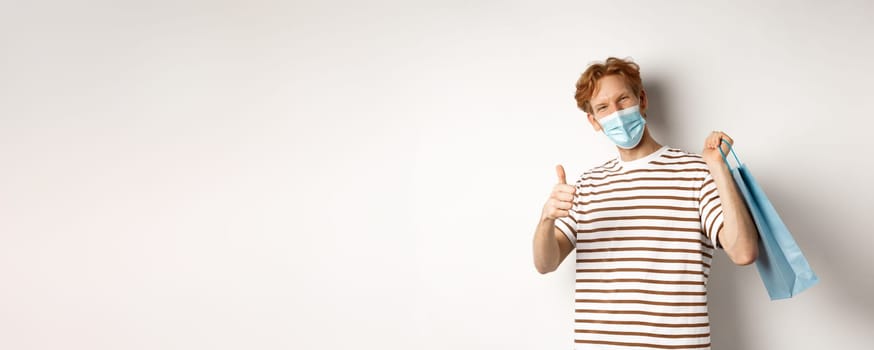 Concept of covid-19 and shopping. Satisfied young man looking pleased after shopping, wearing face mask, showing thumbs-up, recommend store, white background.