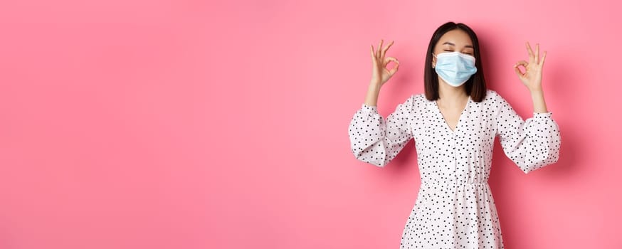 Coronavirus, social distancing and lifestyle concept. Satisfied asian woman in face mask showing okay signs, approve and agree, praising good choice, pink background.