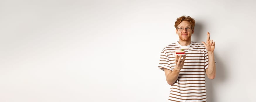 Holidays and celebration concept. Cheerful redhead man in glasses holding birthday cake with candle, cross fingers for good luck and making wish, white background.