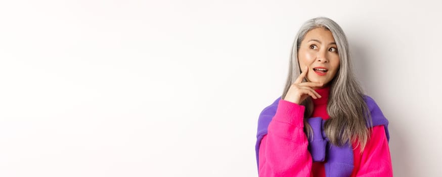 Close up of beautiful asian elderly woman in stylish clothes, making plans, looking aside and thinking, standing over white background.