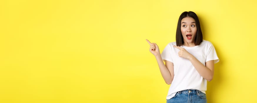 Beauty and fashion concept. Beautiful asian woman in white t-shirt pointing fingers left, standing over yellow background.