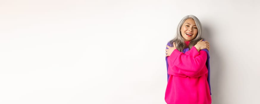 Valentines day and holidays concept. Lovely asian senior woman in pink sweater hugging herself with eyes closed, smiling, standing over white background.