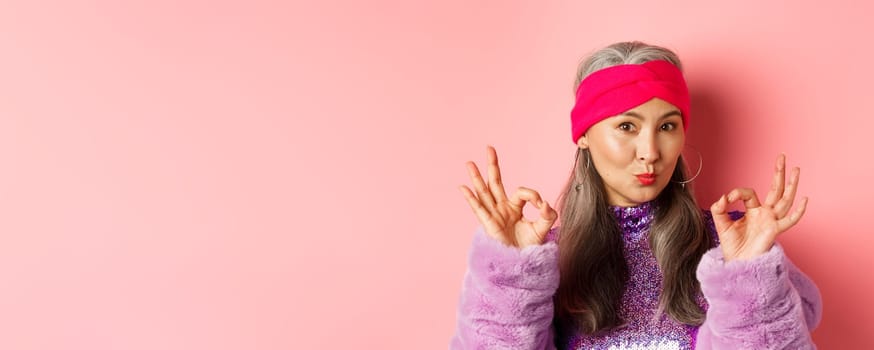 Fashion. Coquettish asian senior woman in trendy headband and faux fur coat, showing okay signs and pucker lips in kiss, praising something perfect, pink background.