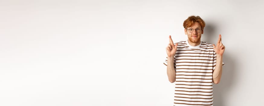 Hopeful young man with red hair and glasses cross fingers for good luck, making wish, standing over white background.