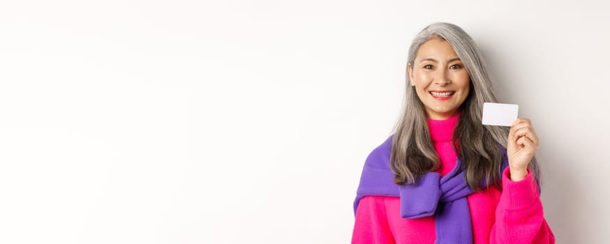 Close-up of beautiful asian senior woman in stylish clothes, smiling and showing plastic credit card of bank, standing over white background.