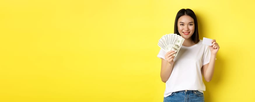 Beautiful asian woman with short dark hair, wearing white t-shirt, showing money in dollars and plastic credit card, standing over yellow background.