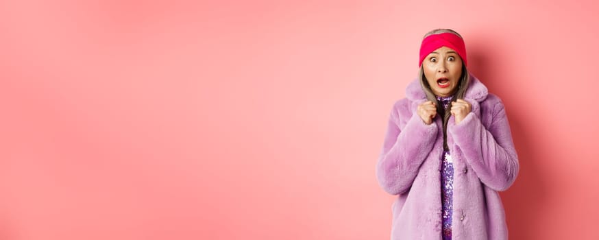 Fashion and shopping concept. Scared elderly woman scream and staring startled at camera, jumping from fear, wearing purple winter coat and headband, pink background.